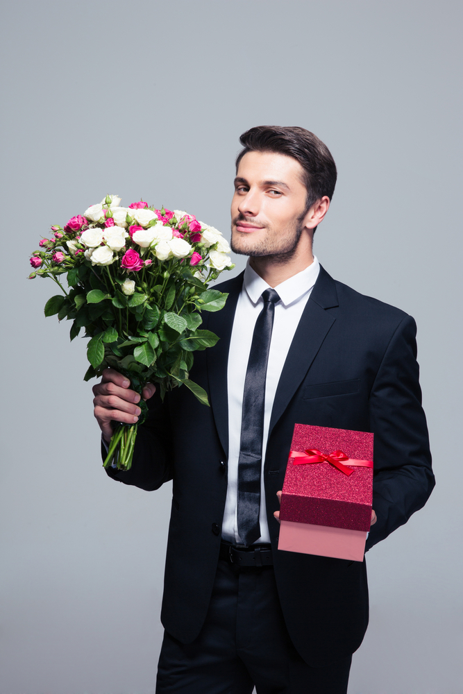 Handsome businessman holding flowers and gift box over gray background. Looking at camera